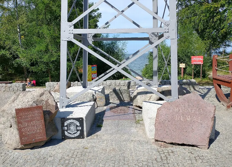 Podstawa krzyża w Pustkowie z kamieniami i tablicami, w tle zejście na plażę
