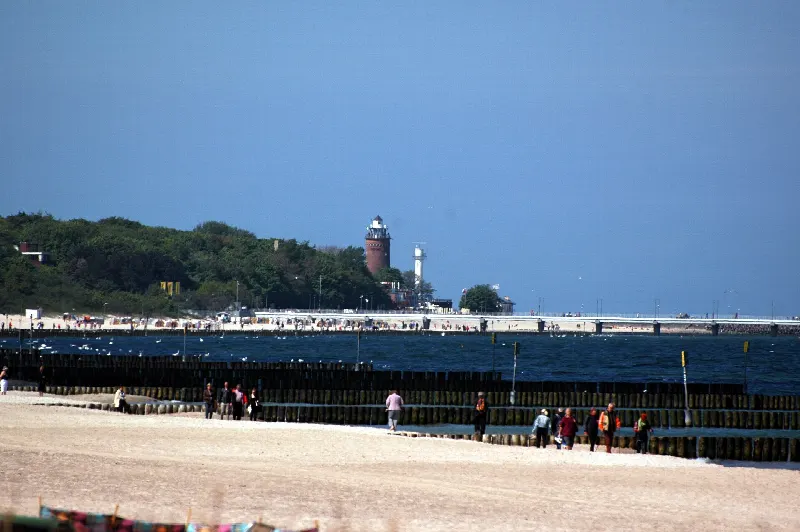 Szeroka plaża wschodnia w Kołobrzegu z drewnianymi ostrogami w morzu i latarnią w tle